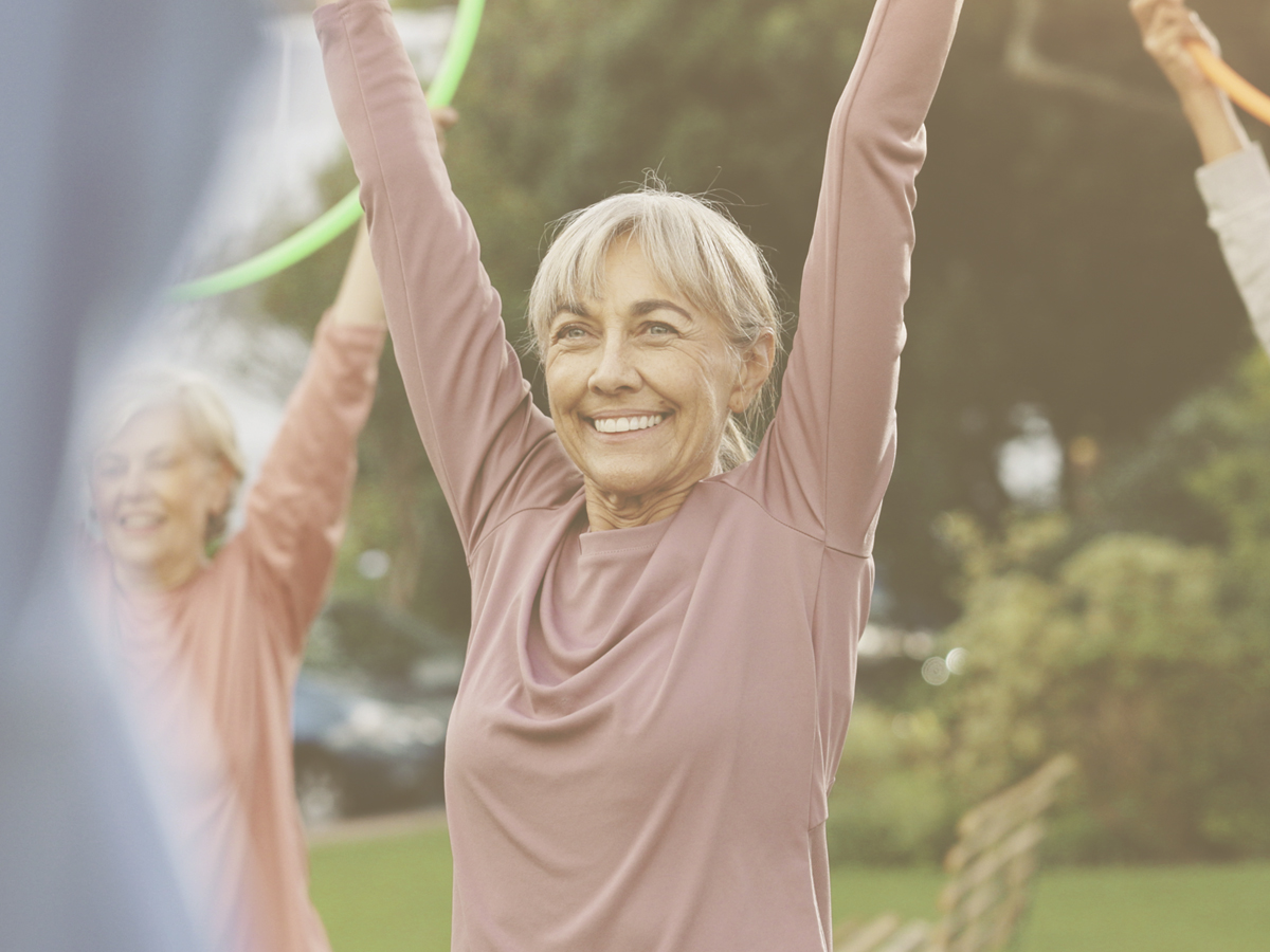 En la imagen vemos a tres señoras mayores sonrientes haciendo ejercicio. Están sujetando un aro con los brazos levantados. La alimentación saludable y la práctica de ejercicio pueden contribuir a prevenir o retrasar la aparición de la hipertensión que se presenta, de manera frecuente, en las personas mayores.