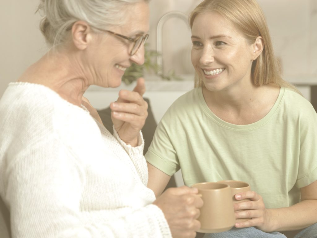 En la imagen vemos a dos mujeres sonrientes sentadas en un sofá tomando una taza de té o café. Una de ellas es una señora mayor y la otra parece ser su hija. Cuando una persona mayor tiene problemas de memoria suele necesitar comprensión. Aprender a hacerlo sin desgastarte tú es interesante para mantener el vínculo emocional.