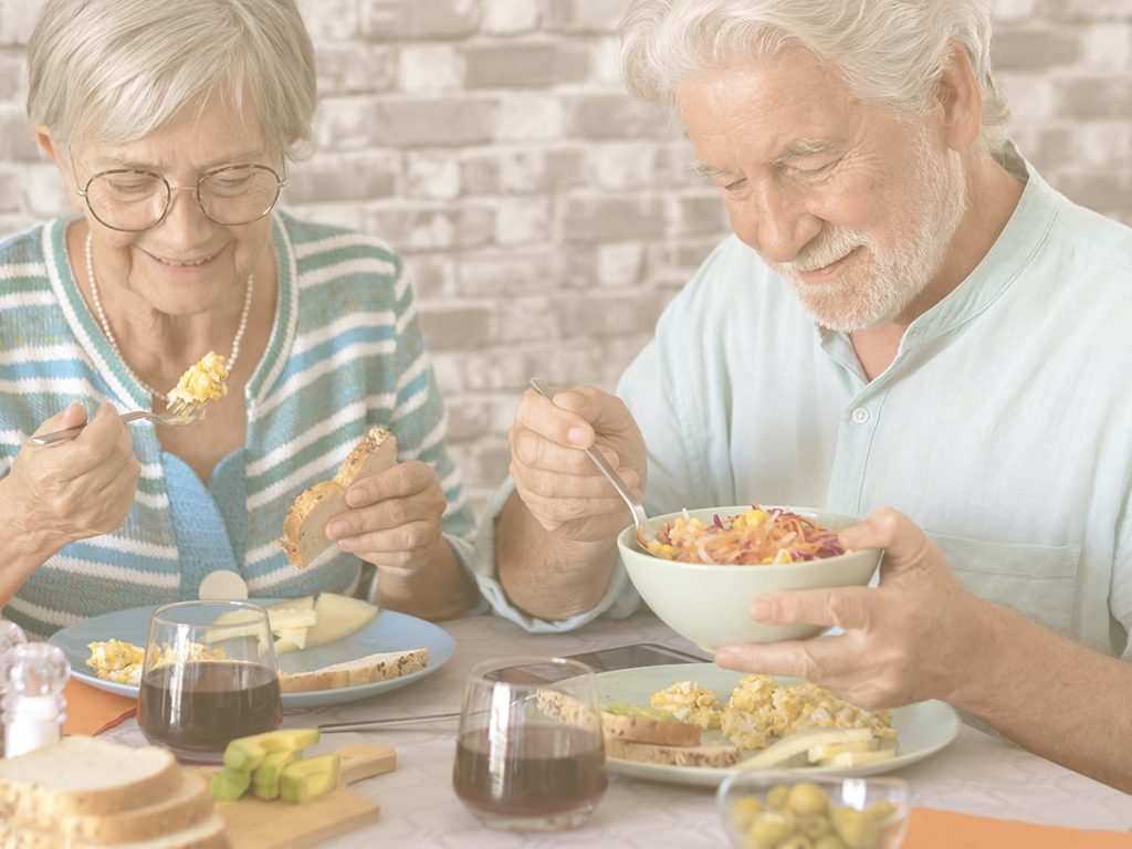 En la imagen, un matrimonio mayor disfruta de un desayuno nutritivo y equilibrado. Ambos sonríen, concentrados en sus alimentos. Una alimentación equilibrada, variada y adaptada a cada momento de la vida es fundamental para garantizar un correcto aporte de nutrientes y para sentirnos bien, tener energía y evitar problemas de salud.