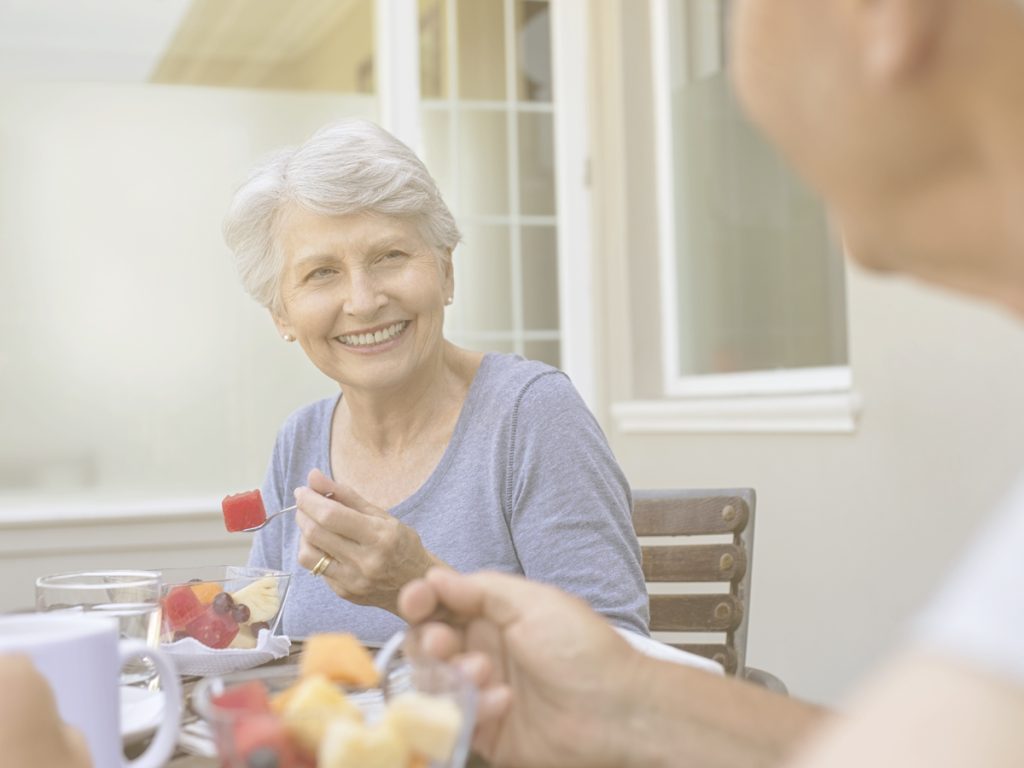 En la imagen vemos a dos personas mayores, una mujer y un hombre que se miran sonrientes sentados a la mesa, comiendo una fruta. Un desequilibro en los niveles de agua corporal y micronutrientes esenciales pueden ser habituales puede comprometer la salud. Por eso, el papel de las personas cuidadoras resulta clave en periodos como el verano, para ayudar a pautar una alimentación adaptada a nuestro familiar.