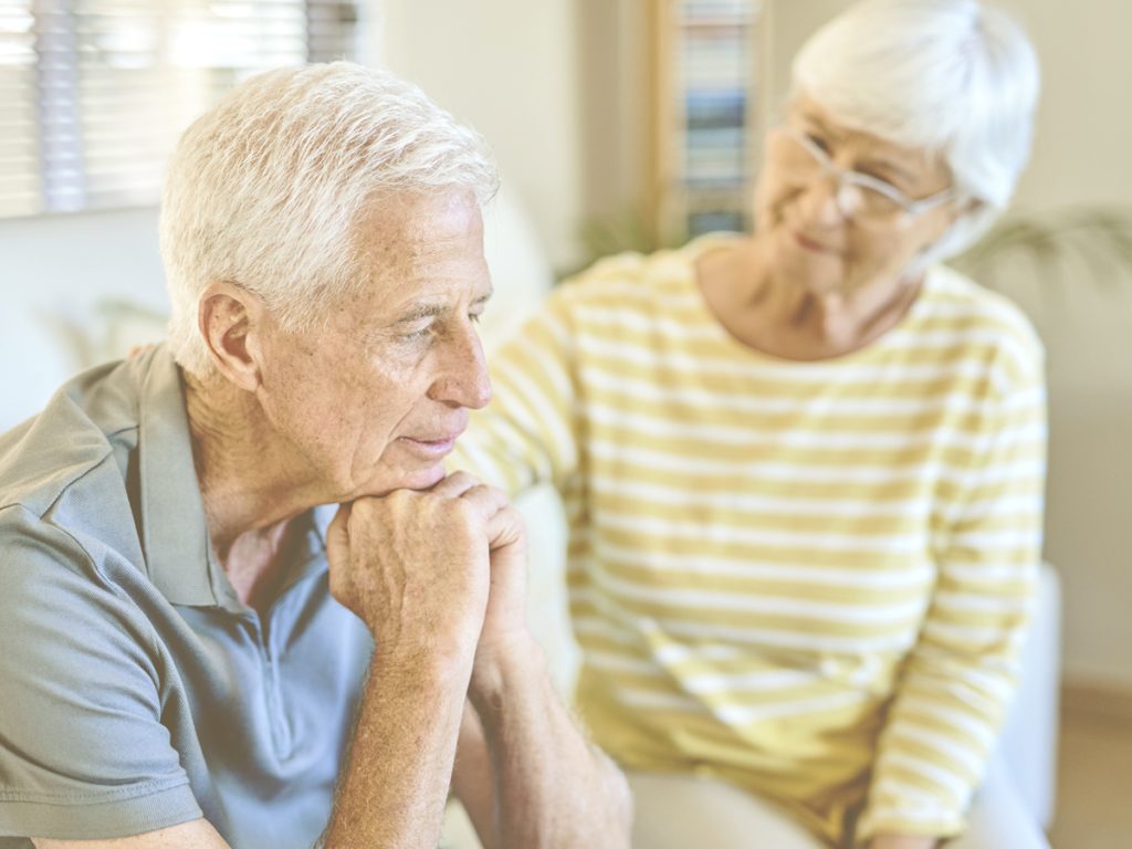 En primer plano, vemos a un anciano sentado en el sofá de su casa, con la cabeza apoyada sobre las manos y gesto de abatimiento o ausencia. Tras él, una mujer mayor, probablemente su esposa, le observa y extiende una mano para brindarle apoyo. A veces, los familiares interpretan las alteraciones psico-conductuales de la persona con demencia como algo voluntario, pero es clave entender que son consecuencia directa de la enfermedad y aprender a identificarlas y gestionarlas.