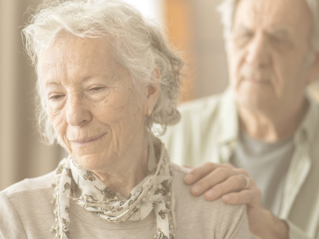 En primer plano, observamos a una anciana con gesto ausente. Tras ella, un hombre mayor, probablemente su marido u otro familiar, posa la mano sobre su hombro, en un claro gesto de apoyo y cariño. El alzhéimer es la enfermedad neurodegenerativa con mayor incidencia a nivel mundial y genera una pérdida progresiva e irreversible de funciones cognitivas. Te indicamos diez señales tempranas para detectar un posible caso de alzhéimer y qué cabe esperar de su evolución.