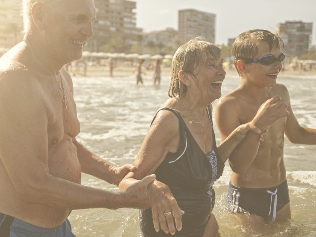 En la iamagen, vemos a tres personas en el mar bañándose y sonriendo. Una mujer y un hombre mayores con el que parece ser su nieto. Todos ellos sonríen mientras se meten en el agua. El señor mayor y el niño sirven como apoyo físico a la mujer. En verano uno de los placeres es disfrutar de baños refrescantes pero, para hacerlo con seguridad, es muy importante que tomemos algunas precauciones.
