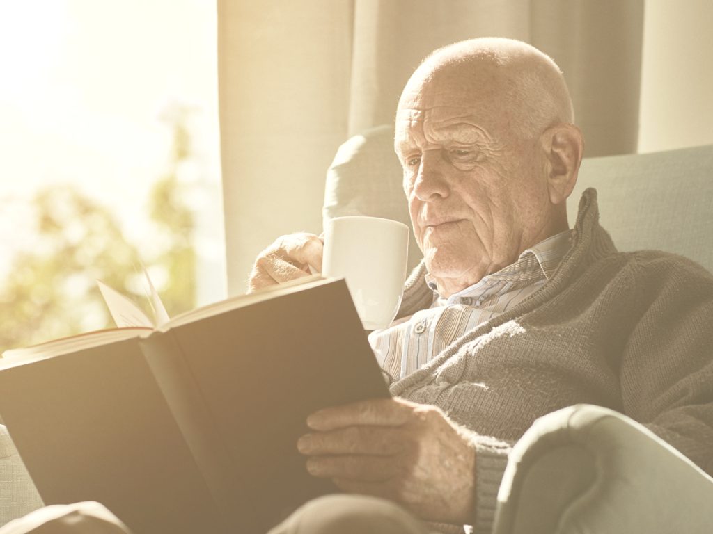 En la imagen, vemos aun hombre mayor sentado en una butaca al lado de una ventana por la que entra mucha luz. El señor está leyendo un libro mientras se toma lo que parece ser una taza de café o un infusión.