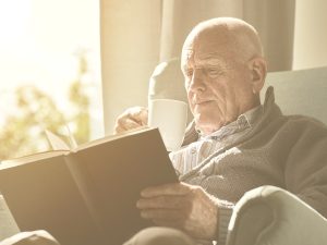 En la imagen, vemos aun hombre mayor sentado en una butaca al lado de una ventana por la que entra mucha luz. El señor está leyendo un libro mientras se toma lo que parece ser una taza de café o un infusión.