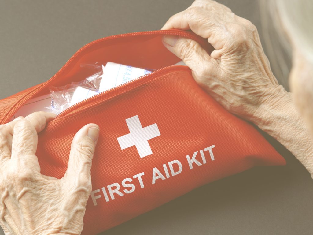 En la imagen, vemos las manos de una mujer mayor sosteniendo una bolsa de tela roja con una cruz blanca y las palabras "First aid kit" -botiquín de primeros auxilios-. A través de la cremallera abierta, intuimos unas gasas o vendas. Contar en casa con un botiquín con artículos de cura y un dispensario con los medicamentos habituales es siempre importante, pero más en el caso de las personas mayores. Además, en su caso, deberemos tener en cuenta ciertos aspectos, para evitar administrarles fármacos sin indicación de su médico que puedan tener contraindicaciones en patologías concretas, o que puedan interaccionar con su medicación habitual, sobre todo cuando se trata de pacientes crónicos o polimedicados.