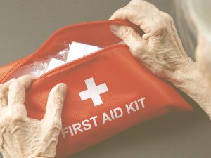 En la imagen, vemos las manos de una mujer mayor sosteniendo una bolsa de tela roja con una cruz blanca y las palabras "First aid kit" -botiquín de primeros auxilios-. A través de la cremallera abierta, intuimos unas gasas o vendas. Contar en casa con un botiquín con artículos de cura y un dispensario con los medicamentos habituales es siempre importante, pero más en el caso de las personas mayores. Además, en su caso, deberemos tener en cuenta ciertos aspectos, para evitar administrarles fármacos sin indicación de su médico que puedan tener contraindicaciones en patologías concretas, o que puedan interaccionar con su medicación habitual, sobre todo cuando se trata de pacientes crónicos o polimedicados.
