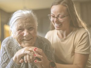 En la imagen vemos a una mujer mayor en primer plano sonriendo y, detrás de ella, la que parece ser su nieta que le sosteniene con cariño y con una sonrisa. El buen trato es la esencia de unos cuidados de calidad. Transciende de atender las necesidades básicas y conlleva reconocer que cada persona es única, con derechos, ideas, prioridades, habilidades y una vida propia.