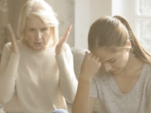 Al fondo de la imagen podemos ver una señora mayor hablando exaltada. En primer plano, una mujer más joven, probablemente su hija, mira hacia abajo con gesto de resignación. Una de las cosas que más desconciertan a quienes cuidan de un familiar es notar que este empieza a tener cambios de humor inesperados: se enfada más, está más irritable, o parece triste sin motivo. Saber identificar y gestionar correctamente estas situaciones es importante en la relación entre personas cuidadoras y cuidadas.