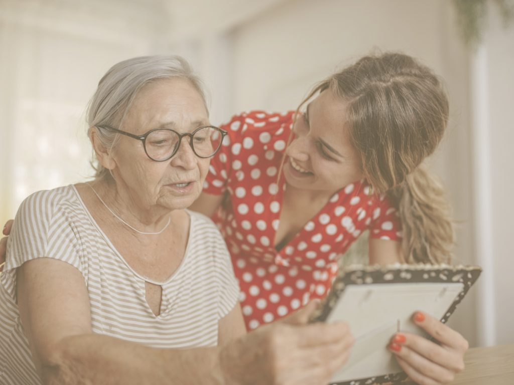 En la imagen vemos a dos mujeres que parecen madre e hija conversando mientras miran una fotografía. En situaciones de deterioro cognitivo leve, la persona conserva la mayor parte de sus capacidades y, en este sentido, es fundamental mantener la conversación con ellas. Buscar imágenes o fotografías pueden servir como apoyo para lograr una comunicación más natural, fluida y que que nos permita estrechar vínculos.