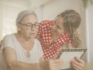 En la imagen vemos a dos mujeres que parecen madre e hija conversando mientras miran una fotografía. En situaciones de deterioro cognitivo leve, la persona conserva la mayor parte de sus capacidades y, en este sentido, es fundamental mantener la conversación con ellas. Buscar imágenes o fotografías pueden servir como apoyo para lograr una comunicación más natural, fluida y que que nos permita estrechar vínculos.