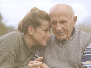 En la imagen, vemos a un hombre anciano sentado en un banco en la calle y, a su lado, a una mujer más joven que podría ser su hija, y que se apoya con cariño en él mientras le coge la mano. Ambos sonríen y transmiten una profunda conexión y ternura.