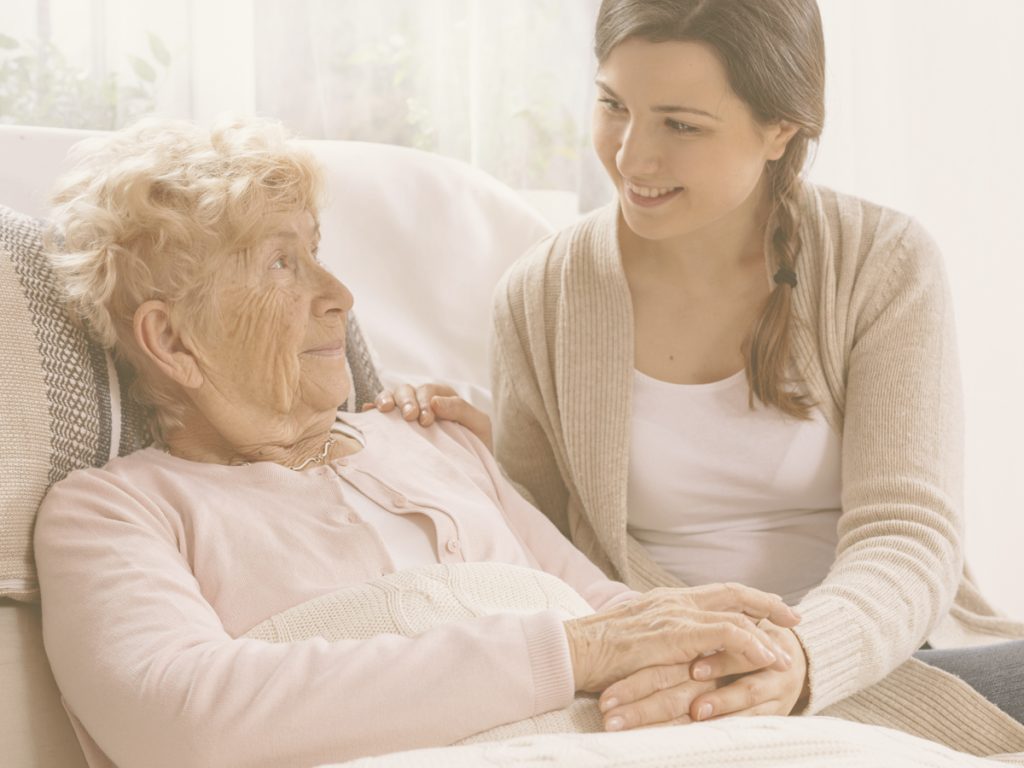 En la imagen, una señora mayor se recuesta en el sofá, mientras una chica joven, probablemente su nieta, le da la mano y le sonríe. Ambas se miran con cariño. Conforme la enfermedad de Parkinson avanza, la autonomía de la persona afectada se va reduciendo, y necesita cada vez más ayuda. Las siguientes recomendaciones pueden facilitarnos la rutina diaria y, sobre todo, aumentar la calidad de vida de nuestro familiar.