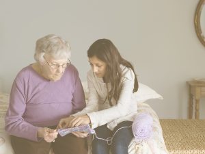Tejiendo vínculos y autonomía en la vejez. En la imagen, una mujer mayor y una joven están sentadas en una cama, compartiendo un momento de tejido. La mujer sostiene la pieza mientras la que, posiblemente, es su nieta, colabora con atención. La escena transmite respeto, conexión intergeneracional y acompañamiento sin infantilización. Fomentar este tipo de relaciones fortalece la autoestima de la persona cuidada y promueve su participación activa en actividades significativas.
