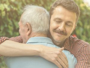 Encontrar un propósito personal al cuidado no significa negar las dificultades, pero dar un sentido a esta experiencia ayuda emocionalmente y refuerza el vínculo con la persona cuidada. En la imagen, vemos a un hombre que abraza a otro más mayor, problablemente su padre, al que vemos de espaldas. El hijo sonríe y transmite una sensación de serenidad.