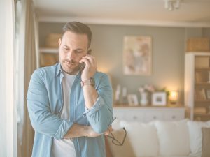 En la imagen, vemos a un hombre de mediana edad en el salón de su casa hablando por el móvil con cara de concentación. Cuidar de un familiar mayor cuando vivimos lejos implica mantener un contacto emocional significativo, organizar apoyos en el entorno más cercano y gestionar emocionalmente los sentimientos de culpa y preocupación.