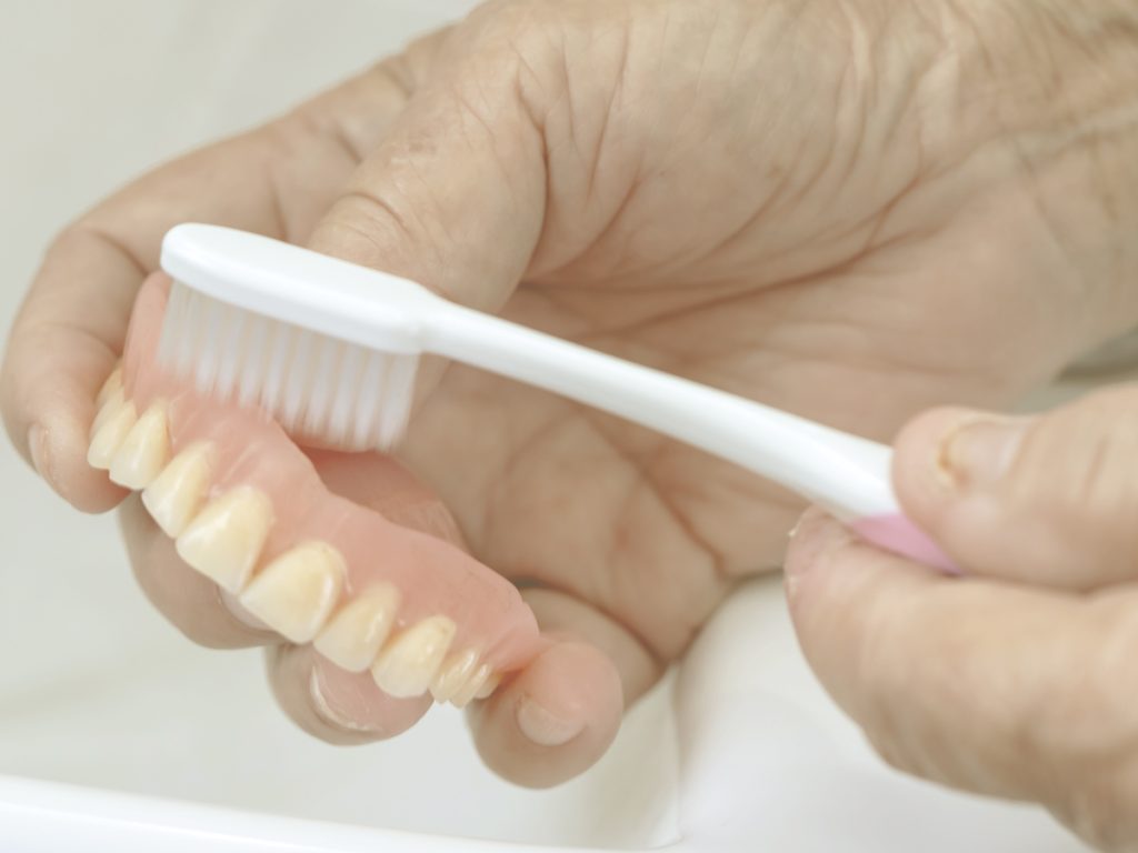 La imagen muestra un primer plano de las manos de una anciana, que sujetan y cepillan cuidadosamente una dentadura postiza. El cuidado de este tipo de prótesis requiere de una higiene diaria para mantenerlas en buen estado y evitar complicaciones bucales.