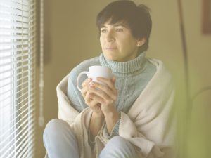 En la imagen vemos a una mujer adulta morena con el pelo negro, en lo que parece el salón de una casa o un dormitorio, envuelta en una manta y sujetando una taza de lo que puede ser un café o una infusión. La señora tiene la mirada perdida en sus pensamientos. En el cuidado de un familiar mayor, tener momentos de descanso y de autocuidado marcan la diferencia para un cuidado de calidad.