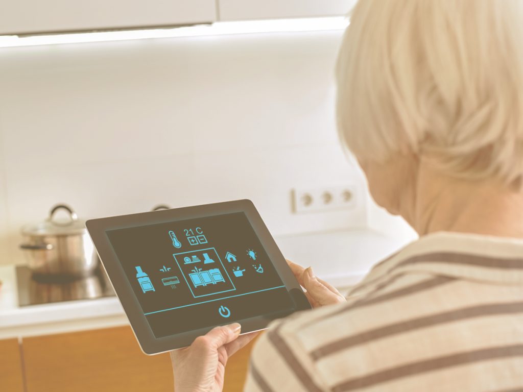 En la imagen vemos a una anciana, sujetando un dispositivo tecnologíco tipo tablet. En la pantalla del aparato aparecen diferentes iconos o símbolos con los que imaginamos que puede controlar algunos dispositivos de su hogar como la cocina, los fuegos, el aire acondicionado, la calefacción, el portal, la puerta de casa, entre otros.