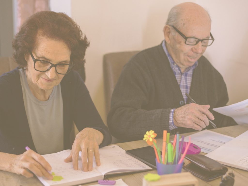 En la imagen vemos a un matrimonio de ancianos sentados en una mesa en la que hay papeles, libretas y bolis de colores. Ambos están escribiendo en algunos de los papeles. El cálculo es una de las aptitudes necesarias para desenvolvernos en el día a día.