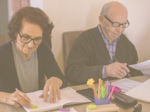 En la imagen vemos a un matrimonio de ancianos sentados en una mesa en la que hay papeles, libretas y bolis de colores. Ambos están escribiendo en algunos de los papeles. El cálculo es una de las aptitudes necesarias para desenvolvernos en el día a día.
