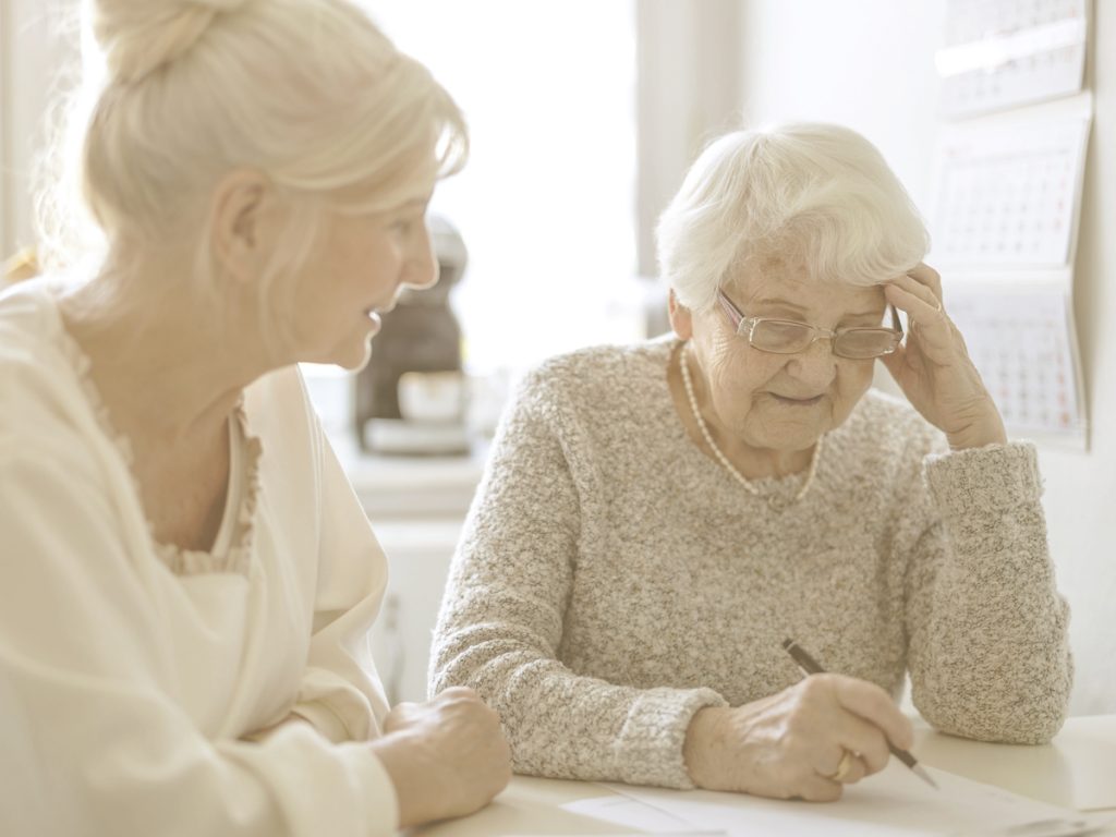 En la imagen vemos a una anciana sentada en la mesa de su cocina, concentrada en realizar una ficha de ejercicios que tiene delante. Junto a ella vemos a una mujer de mediana edad, posiblemente su hija, que la observa y parece ayudarla. Las habilidades cognitivas, como la orientación, pueden verse mermadas con la edad, pero pueden trabajarse con ejercicios sencillos.