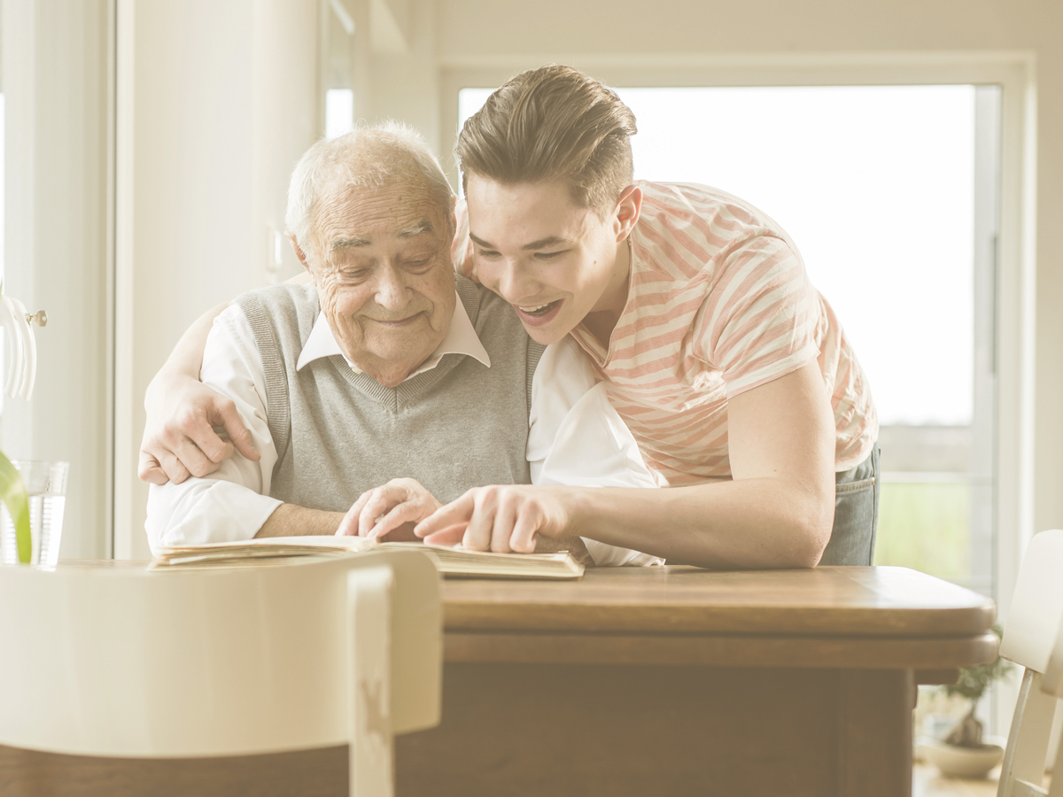 En la imagen vemos a un abuelo con su nieto en el salón de una casa. El nieto abraza al abuelo con cariño mientras ambos miran un libro u album. Ejercitar la memoria es especialmente importante en personas mayores ya que es una de la habilidades cognitivas fundamentales para el día a día y, con la edad, la memoria disminuye.