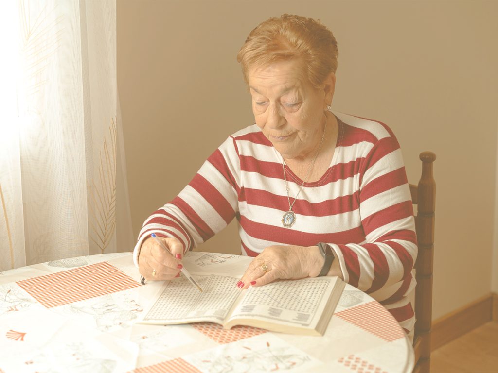 En la imagen, vemos a una señora mayor, sentada en una mesa en su casa, concentrada en resolver crucigramas en un cuaderno de pasatiempos. Poner atención a la hora de recibir y procesar la información es clave para almacenar y recordar con facilidad esos datos, y continuar entrenando esta habilidad, también en la tercera edad, nos ayudará a mantener la agilidad mental.