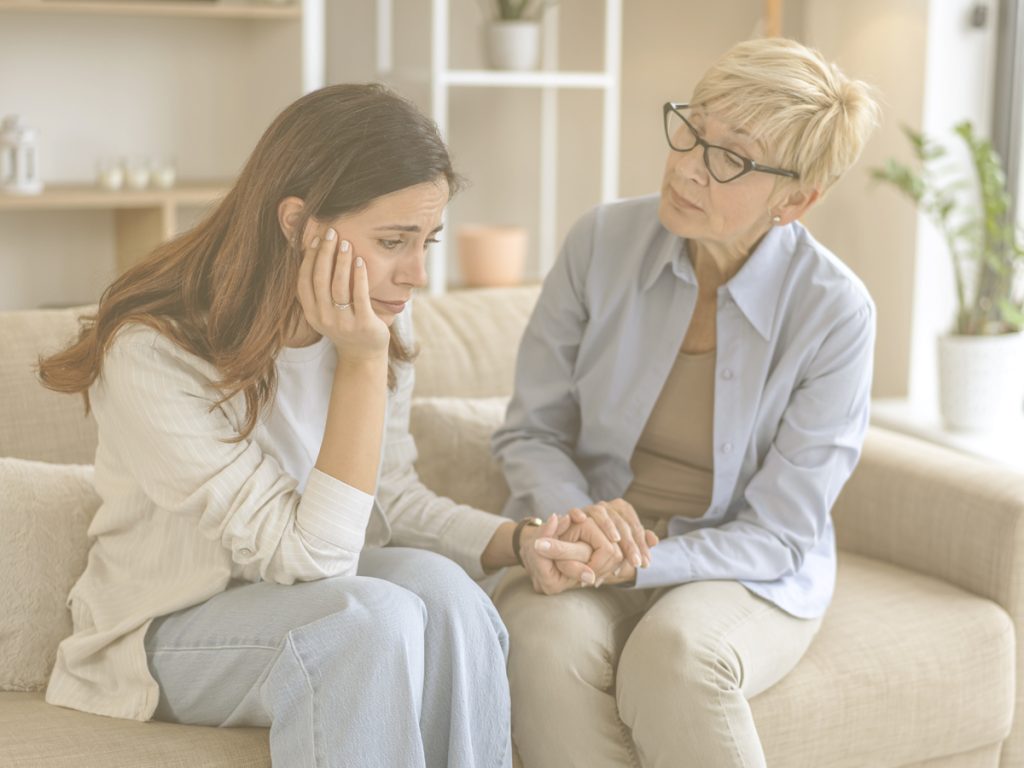En la imagen vemos a dos mujeres, una mayor y otra de mediana edad, sentadas en un sillón en un salón de una casa. La mujer mayor sostiene con sus dos manos una de las manos de la otra mujer que parece tener un gesto de preocupación.