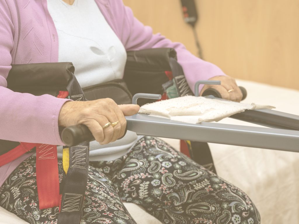 En la imagen vemos las piernas de una mujer mayor que, sentada en la cama, agarra el asidero de una grúa. Existen distintos tipos de grúas que pueden servirnos de ayuda en el hogar: las de bipedestación, y las de tipo cigüeña. Además, también hay otros dispositivos como elevadores de asa, protecciones laterales de la cama y estribos de sujeción que pueden resultar de utilidad.