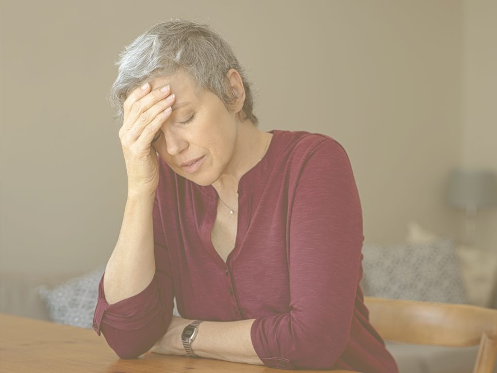 En la imagen, una mujer de mediana edad con gesto preocupado se sienta en el suelo, a los pies de su cama. Se la ve preocupada, con gesto reflexivo e, incluso, triste. Aprender a gestionar el estrés y la ansiedad en situaciones de cuidado, sobre todo cuando estas se prolongan en el tiempo, y dedicar atención al autocuidado resulta clave para el bienestar emocional de las personas cuidadoras.