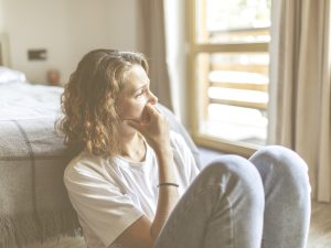 En la imagen, una mujer de mediana edad con gesto preocupado se sienta en el suelo, a los pies de su cama. Se la ve preocupada, con gesto reflexivo e, incluso, triste. Aprender a gestionar el estrés y la ansiedad en situaciones de cuidado, sobre todo cuando estas se prolongan en el tiempo, y dedicar atención al autocuidado resulta clave para el bienestar emocional de las personas cuidadoras.
