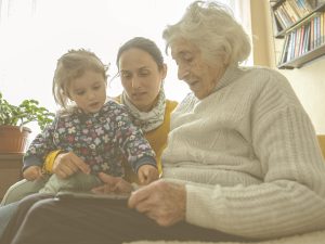 En la imagen, vemos a una mujer de mediana edad sentada en un sofá o una cama con una anciana, que podría ser su madre, y una niña de cuatro o cinco años, probablemente, su hija. Las tres parecen fijarse en la tablet que la abuela tiene en su regazo. La conciliación entre el cuidado de nuestros mayores y nuestros hijos e hijas es una realidad cada vez más frecuente en nuestra sociedad, una situación que se aplica a la denominada "generación sandwich".