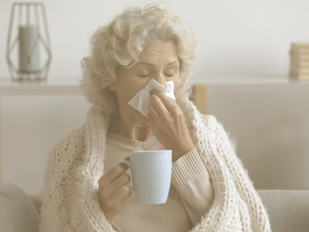 En la imagen, observamos a una mujer mayor sentada en el sofá de su casa que, abrigada con un chal de lana, se suena la nariz con un pañuelo desechable. En la otra mano, sostiene una taza de lo que podemos imaginar que será una infusión caliente. Las infecciones respiratorias como la gripe entrañan mayor riesgo para las personas mayores, dado que el sistema inmunitario se debilita con la edad y que pueden tener otras enfermedades crónicas que aumentan la probabilidad de complicaciones.