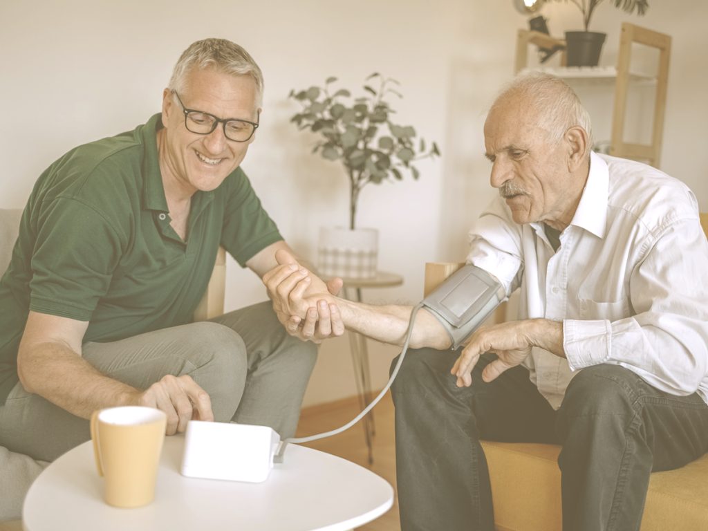 En la imagen vemos a dos hombres en un salón sentados en unos sofás. Uno de ellos es anciano y el otro de mediana edad. El más joven está tomando la tensión al hombre más mayor. Cuando cuidamos a una persona mayor, hemos de estar atentos a una posible hipertensión de la persona a nuestro cargo para detectarla cuanto antes y prevenir sus consecuencias. Para ello, es importante controlar de manera periódica la tesión arterial.