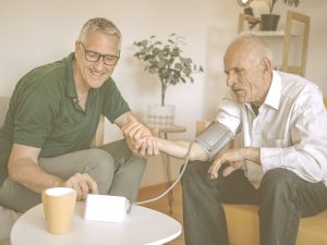 En la imagen vemos a dos hombres en un salón sentados en unos sofás. Uno de ellos es anciano y el otro de mediana edad. El más joven está tomando la tensión al hombre más mayor. Cuando cuidamos a una persona mayor, hemos de estar atentos a una posible hipertensión de la persona a nuestro cargo para detectarla cuanto antes y prevenir sus consecuencias. Para ello, es importante controlar de manera periódica la tesión arterial.