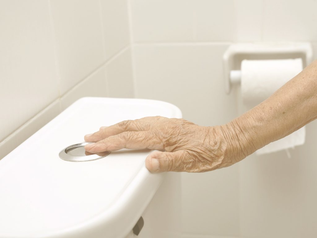 En la imagen vemos la mano de un anciano o anciana pulsando el botón de la taza de un water para tirar de la cadena en el baño de una casa.