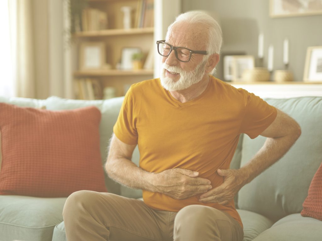 En la imagen, vemos a un señor mayor sentado en el sofá de su casa, con las manos en el costado y gesto de dolor. Las infecciones de orina pueden prevenirse con una correcta hidratación, alimentación y rutinas de aseo.