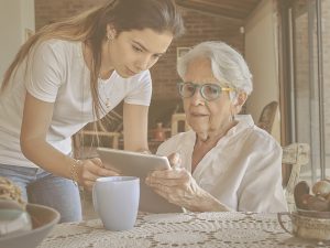 ¿Es positivo implicar en el cuidado a las generaciones más jóvenes? En la imagen, una mujer mayor y una joven están sentadas en una mesa. La joven se inclina para mostrarle a la mujer cómo usar una tablet, mientras ambas observan la pantalla con atención. La escena refleja un acompañamiento respetuoso y paciente, donde la tecnología se convierte en un puente entre generaciones. Incluir a personas jóvenes en el cuidado puede fortalecer vínculos familiares y promover una mirada más inclusiva y empática hacia el envejecimiento.