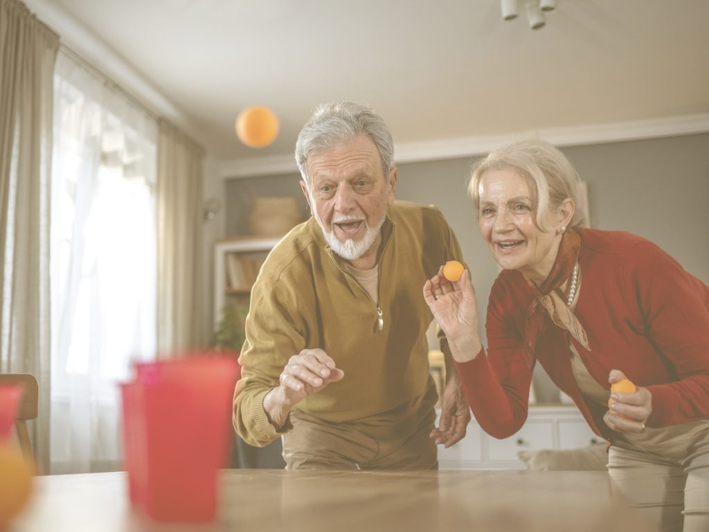 En la imagen vamos a dos personas de mediana edad jugando en el salón de su casa con gesto divertido. Encima de la mesa del salón tienen varios vasos de plástico en los que están intentando canastar unas pelotas pequeñas tipo ping pong. Que los ancianos realicen ejercicio físico de manera habitual es fundamental para evitar problemas de movilidad y continuar siendo autónomos. También ayuda a mantener la salud cardiovascular y a la disminución de la aparición de enfermedades. Y, por último, es muy beneficioso a nivel emocional y psicológico.