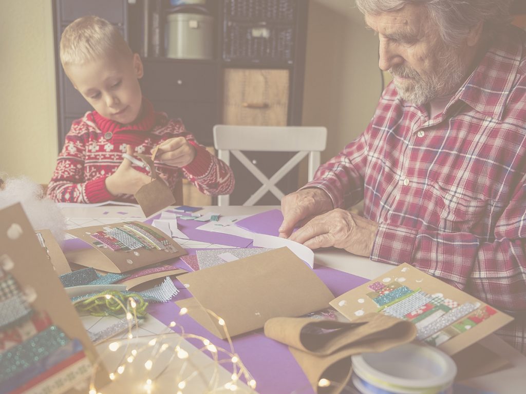 En la imagen, vemos a un niño de unos siete años y a su abuelo, sentados en la mesa de su casa. Esta está cubierta por distintos materiales de manualidades, como cartulinas, cintas y luces, y ambos están concentrados en la elaboración de unas tarjetas de Navidad artesanales. Las manualidades sirven a mayores y niños para estimular habilidades cognitivas como la concentración y la atención, además de entrenar la motricidad fina. Además, son una excelente opción para compartir tiempo en familia.