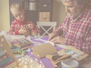 En la imagen, vemos a un niño de unos siete años y a su abuelo, sentados en la mesa de su casa. Esta está cubierta por distintos materiales de manualidades, como cartulinas, cintas y luces, y ambos están concentrados en la elaboración de unas tarjetas de Navidad artesanales. Las manualidades sirven a mayores y niños para estimular habilidades cognitivas como la concentración y la atención, además de entrenar la motricidad fina. Además, son una excelente opción para compartir tiempo en familia.