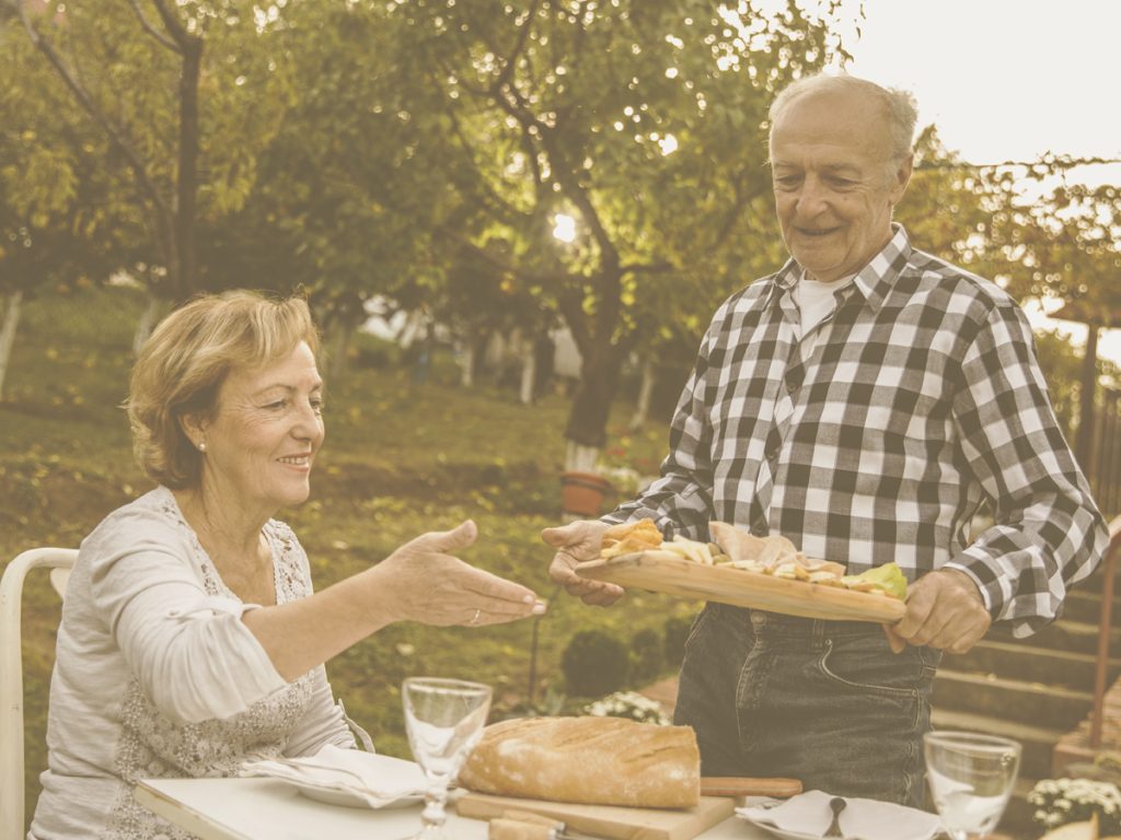 En la imagen vemos a una señora y un señor mayor preparados para comer en un jardín. La señora está sentada y el señor está colocando una bandeja en la mesa que ya está preparada con el mantel, los platos, cubiertos y copas. Una correcta alimentación durante todo el año es clave para mantener la salud de los ancianos. En primavera, la exposición reponsable al sol, decansar, hidratarse y consumir alimentos de temporada, favorecen el bienestar de las personas mayores en esta época del año.