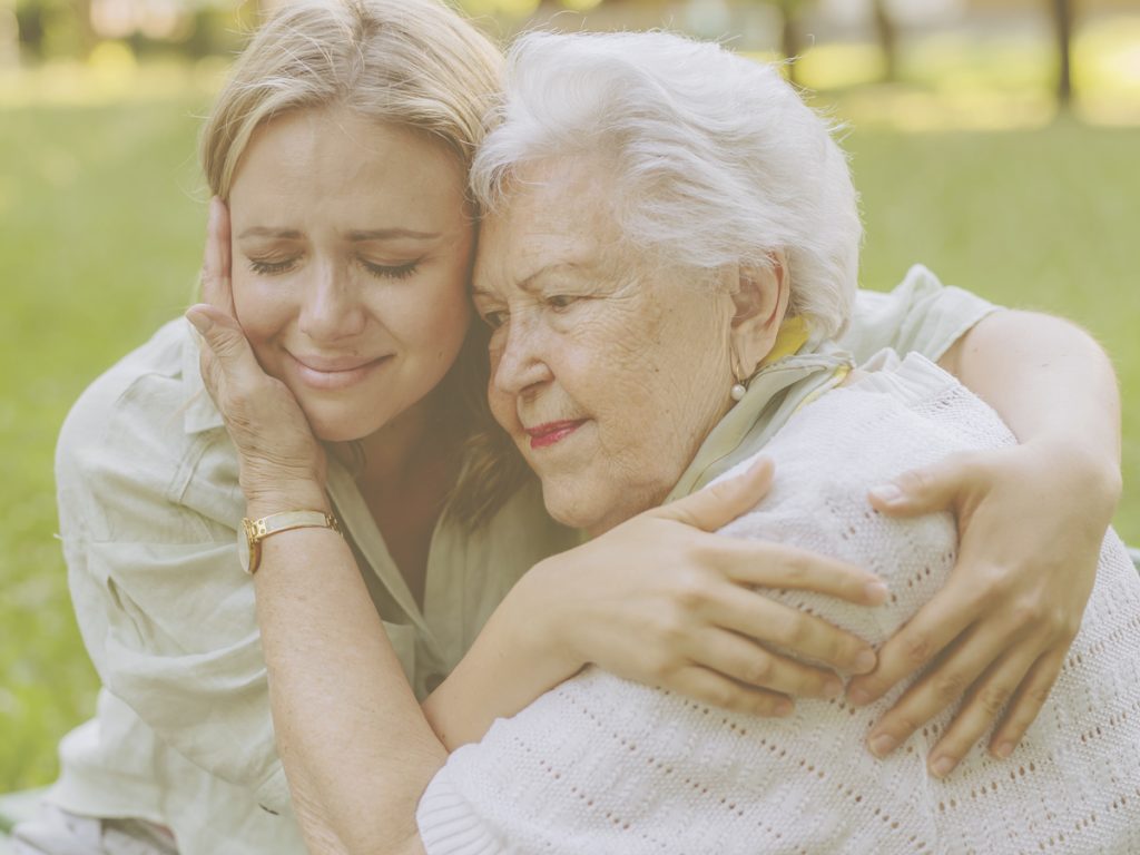 En la imagen, vemos a dos mujeres, una chica joven y una señora mayor, sentadas en un banco de un jardín dándose un abrazo. La joven parece tener cara de tristeza y preocupación y la anciana le sostiene la cara con la mano en un gesto que parece de consuelo. En el cuidado de un familiar, el miedo al futuro es una de las emociones más habituales y se puede intentar gestionar mejor con algunas claves como El miedo al futuro es una de las emociones más comunes durante el cuidado de un familiar