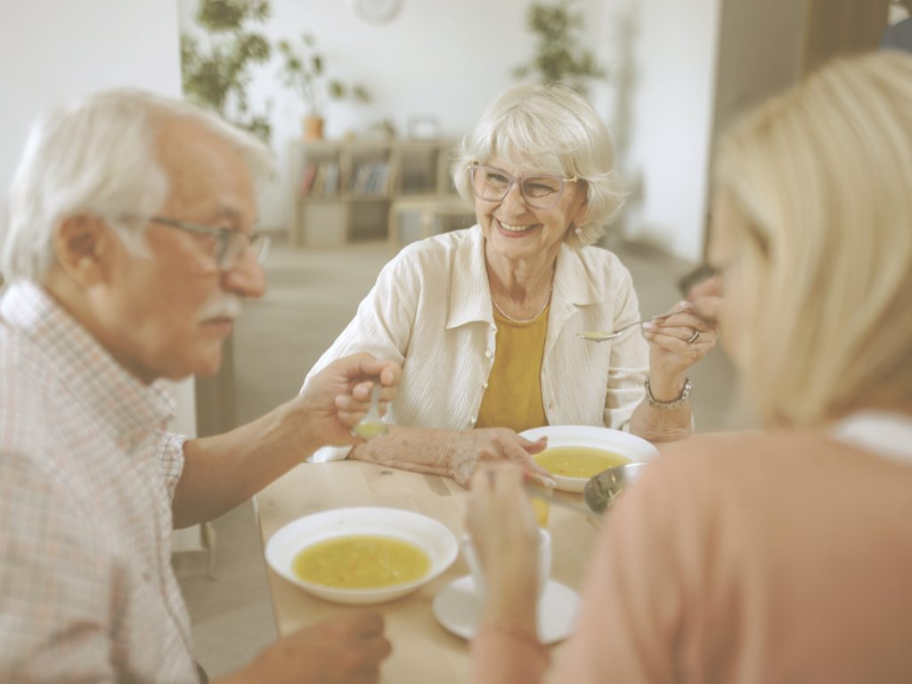 En la imagen vemos a tres personas mayores sentadas alrededor de una mesa mientras comen juntas. Una mujer sonríe mientras conversa con los demás, en un ambiente distendido y cercano. Compartir las comidas con otras personas puede favorecer la socialización, ayudar a mantener rutinas y convertir la alimentación en un momento agradable que contribuya al bienestar emocional de las personas mayores.