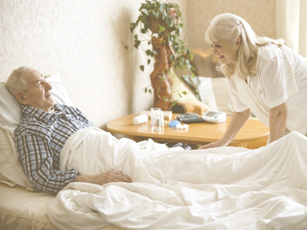 En la imagen, vemos a un señor mayor tumbado boca arriba con gesto sonriente en una cama y a una señora mayor inclinada ayudando a que la persona que está encamada tenga una posición más cómoda y adecuada. Los cambios de postura en la cama son fundamentales para la salud, ya que, entre otros beneficios, reducen los puntos de presión sobre el cuerpo y mejoran la circulación sanguínea.