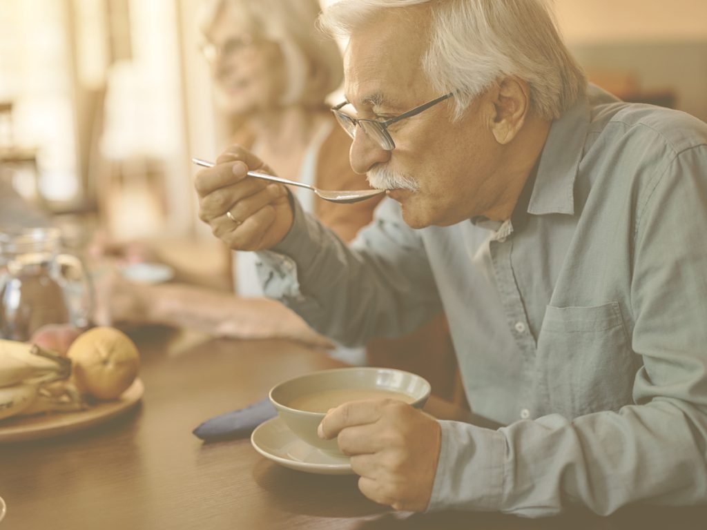 En la imagen, vemos a un señor mayor de pelo cano, de aspecto saludable que, sentado a la mesa con su familia, se lleva una cucharada de sopa a la boca. En los meses de invierno, es conveniente adaptar la alimentación, sobre todo en el caso de las personas mayores, para garantizar un correcto aporte de nutrientes, así como una adecuada hidratación y tolerancia digestiva. Sopas y caldos, legumbres y guisos tradicionales resultan una excelente opción.