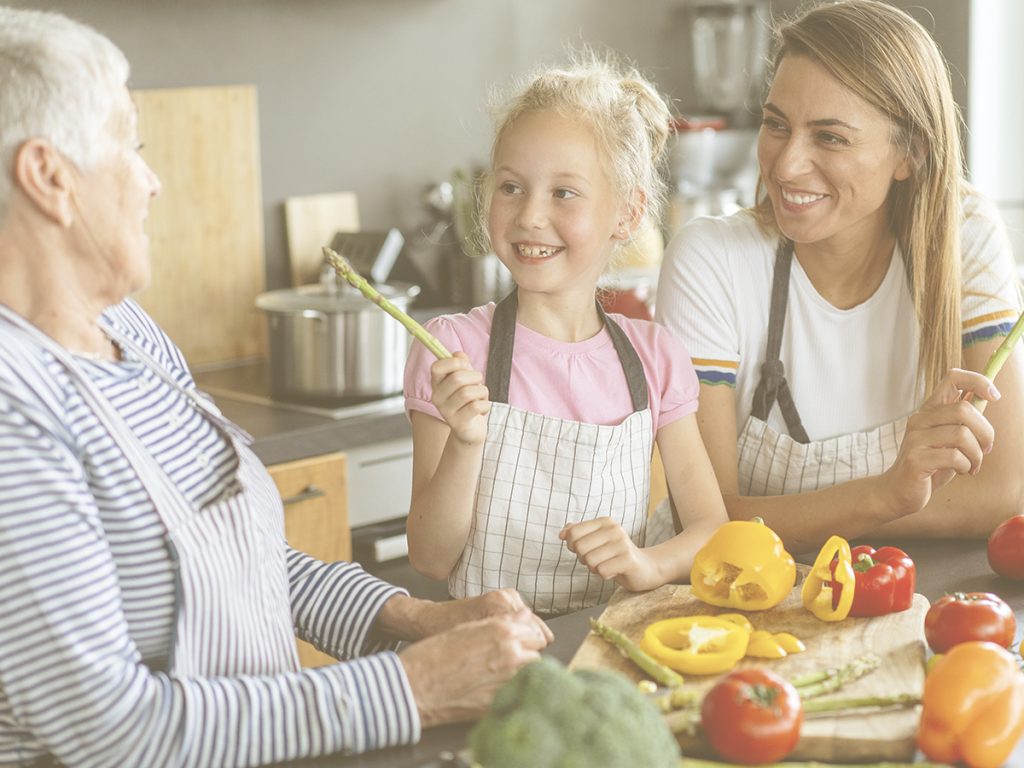 En la imagen, vemos a una familia apoyada en la encimera de su cocina, donde podemos identificar distintas verduras y hortalizas: brócoli, pimientos, tomates de distinto tipo... En el centro, una niña sonríe mientras le muestra un espárrago a su abuela. Junto a ellas, la madre sonríe. La Navidad, con tantas comidas y cenas previstas, puede ser una buena ocasión para cocinar juntos. Ofrecemos una receta de carne y otra de pescado para comer sabroso y saludable en estas fechas. Opciones ricas en proteínas y de fácil masticación y digestión para personas mayores.