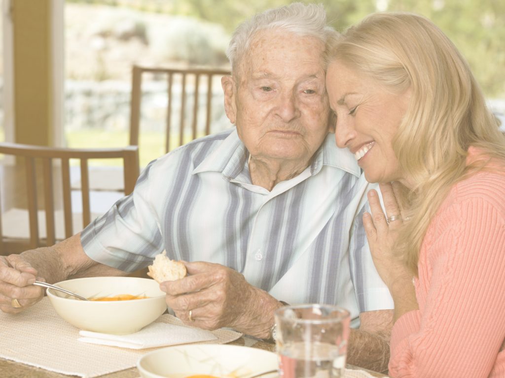 En la imagen vemos a un señor mayor y a una mujer de mediana edad sentados a la mesa en una terraza en un día soleado. El hombre está comiendo y la mujer le abraza en un gesto de cariño. En la época estival y cuando las temperaturas se incrementan de manera constante, el cuerpo y el funcionamiento de las personas mayores se altera. El estreñimiento es uno de los malestares más comunes en este periodo. Una dieta rica que aporte hidratación y fibra puede ayudar a contrarrestar esta tendencia.