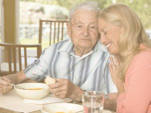 En la imagen vemos a un señor mayor y a una mujer de mediana edad sentados a la mesa en una terraza en un día soleado. El hombre está comiendo y la mujer le abraza en un gesto de cariño. En la época estival y cuando las temperaturas se incrementan de manera constante, el cuerpo y el funcionamiento de las personas mayores se altera. El estreñimiento es uno de los malestares más comunes en este periodo. Una dieta rica que aporte hidratación y fibra puede ayudar a contrarrestar esta tendencia.